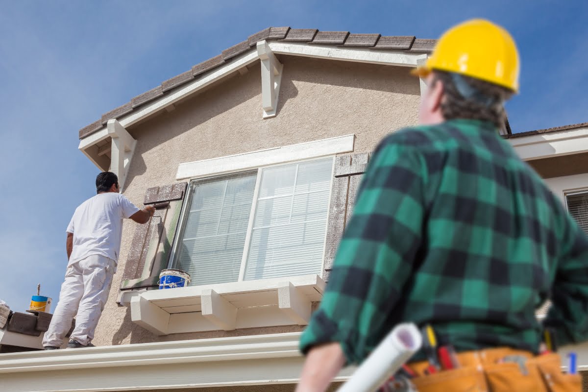 A crm for painting contractors has a painter inworking attire paints the exterior of a house from a ladder while another individual in a yellow hard hat observes from below.