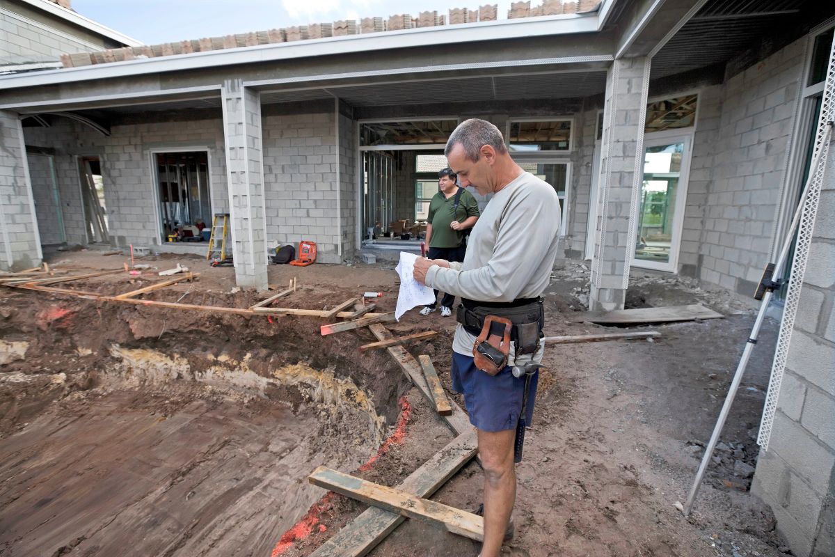 A construction worker reviews a document at a residential construction site with a partially built cinder block structure and dug-out area, possibly for pool installation. A second worker stands in the background, perhaps discussing details using pool construction software.