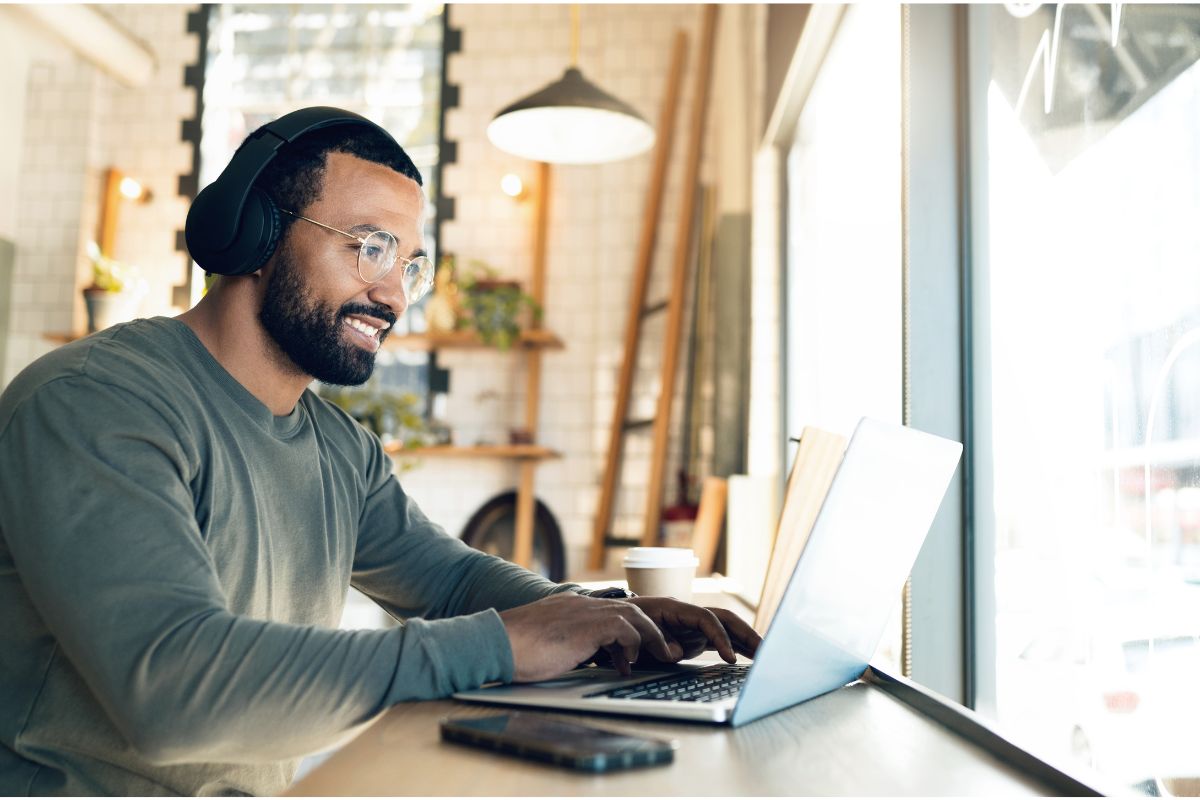 A man wearing headphones and glasses types on a laptop at a cafe table, engaging in remote work. A smartphone and a coffee cup sit beside him.