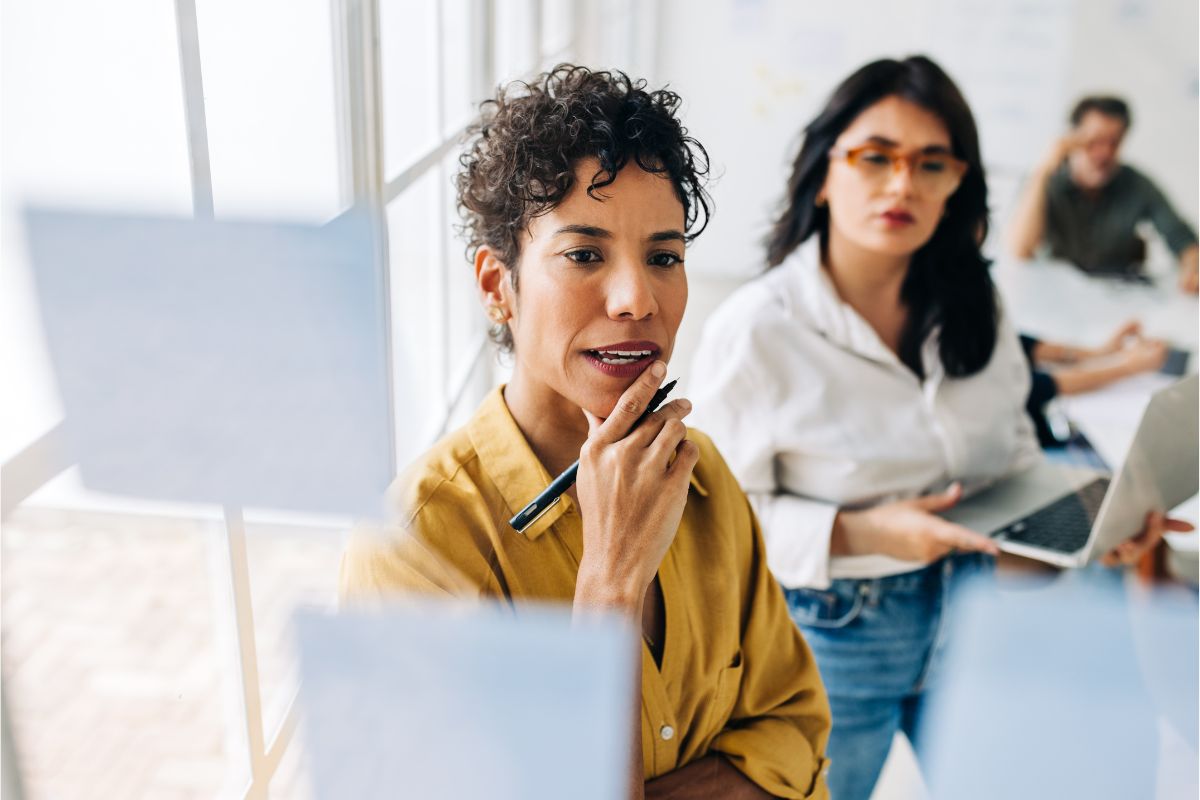 Two women in an office setting brainstorm while looking at sticky notes on a glass wall; one holds a pen to her chin, and the other stands with a laptop, discussing how remote work has influenced their strategies.