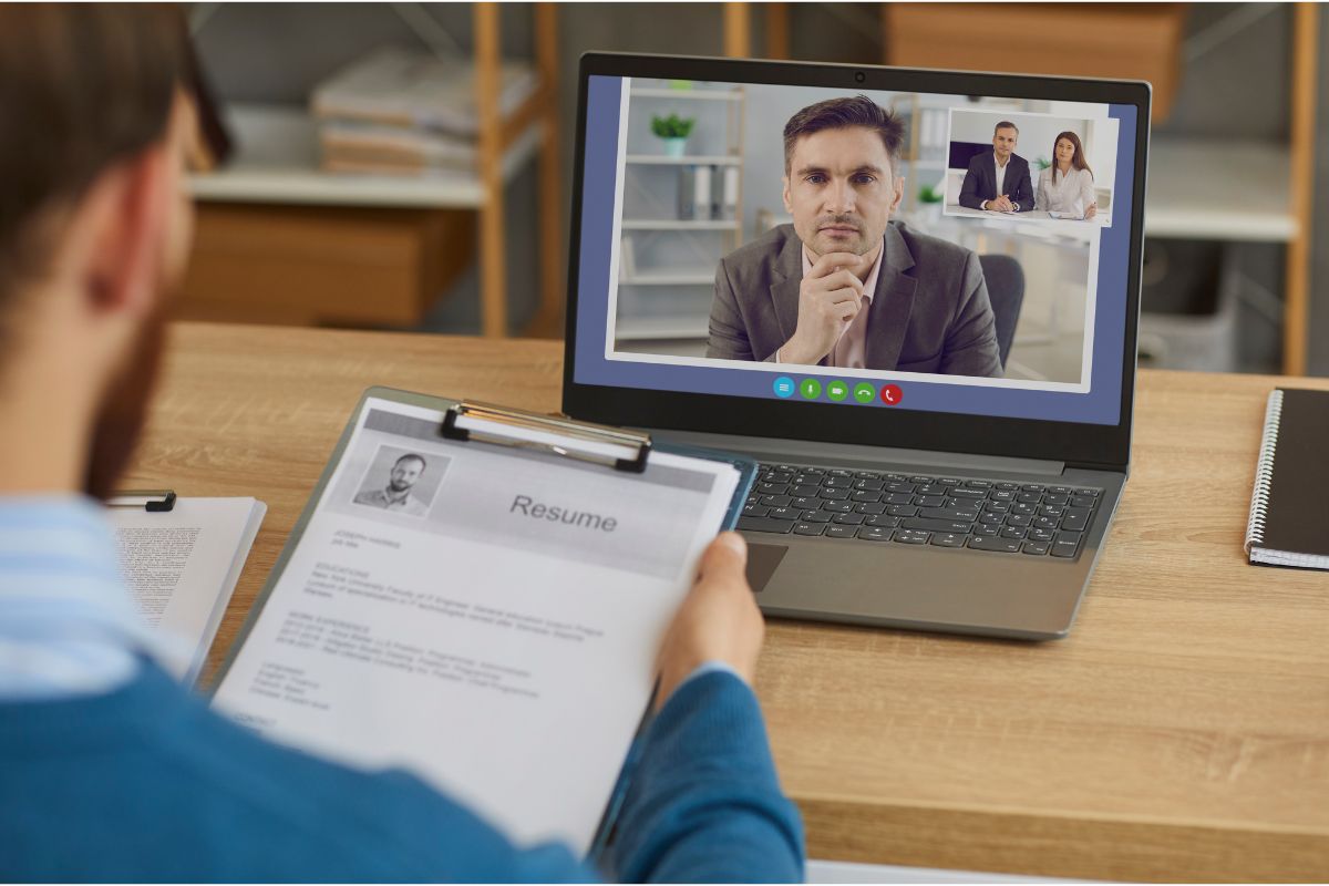 A person holds a resume while participating in a video call interview on a laptop with two individuals visible on the screen, exemplifying the seamless integration of remote work into modern hiring practices.