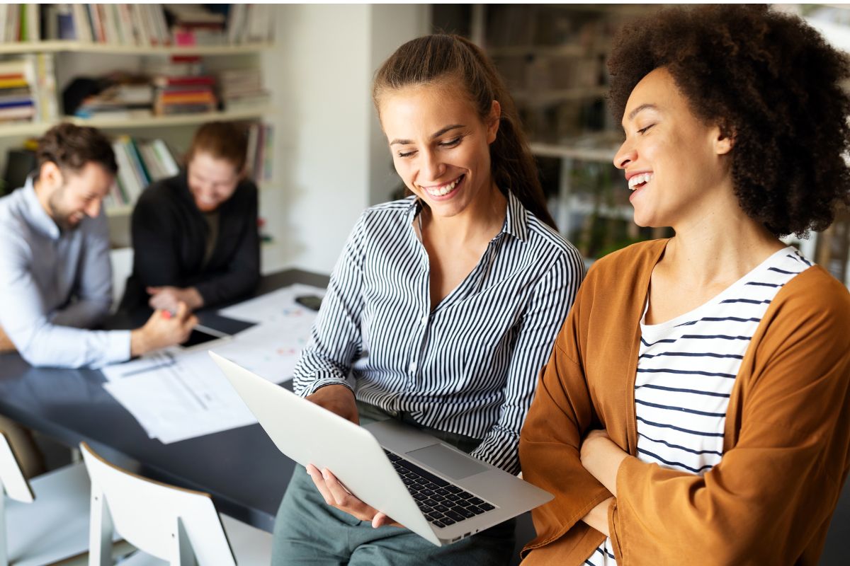 Two women are smiling while looking at a laptop in an office, possibly discussing robots.txt for SEO strategies. Two people are working at a table in the background, and bookshelves are visible behind them.