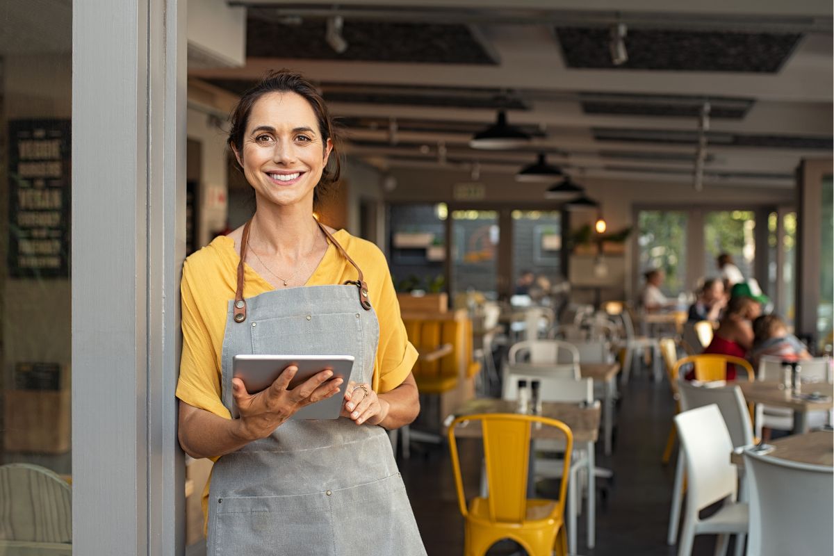 A smiling woman in a yellow shirt and gray apron holds a tablet while standing in a modern, brightly-lit café with customers in the background, seamlessly managing SEO for multiple locations.