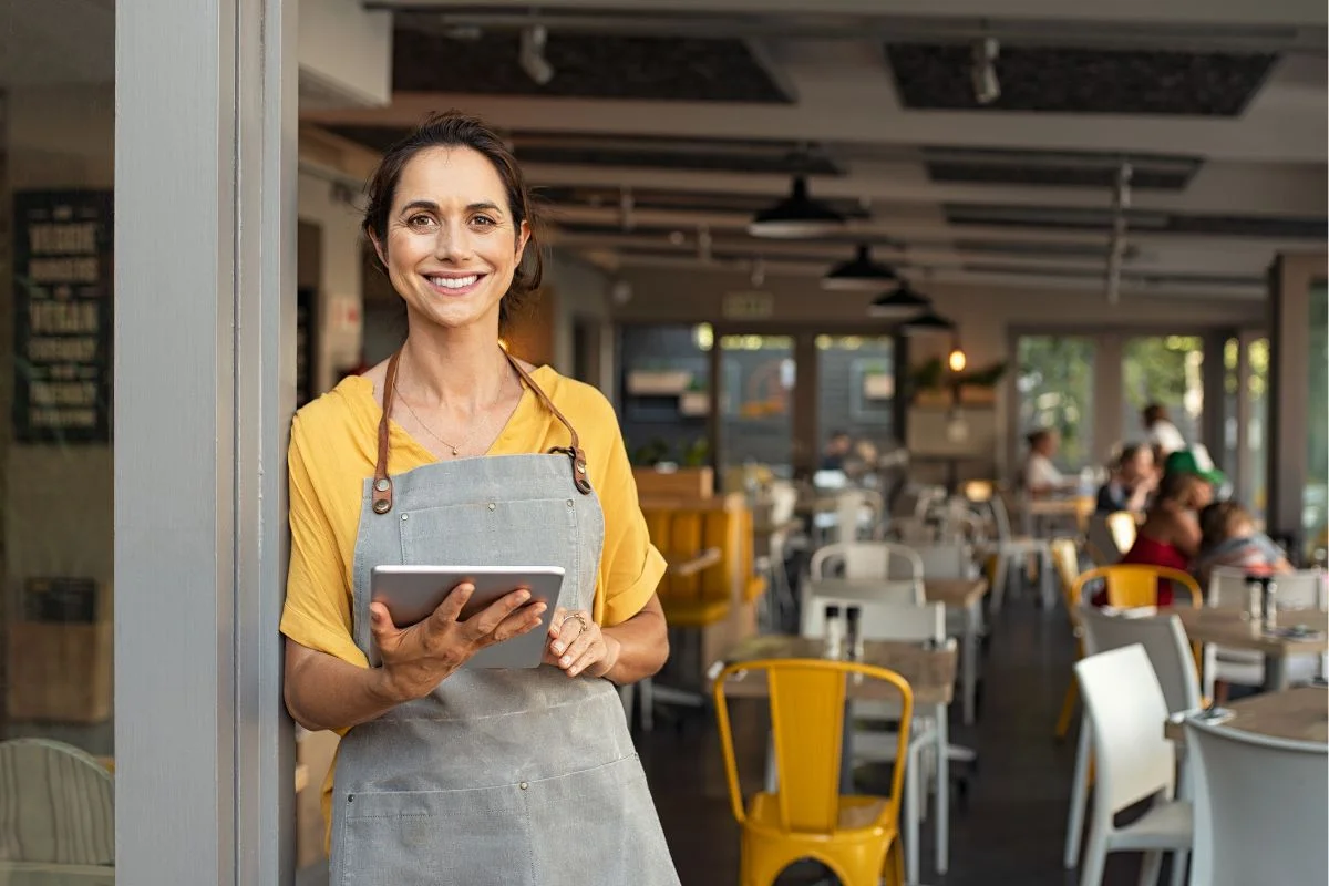 A smiling woman in a yellow shirt and gray apron holds a tablet while standing in a modern, brightly-lit café with customers in the background, seamlessly managing SEO for multiple locations.