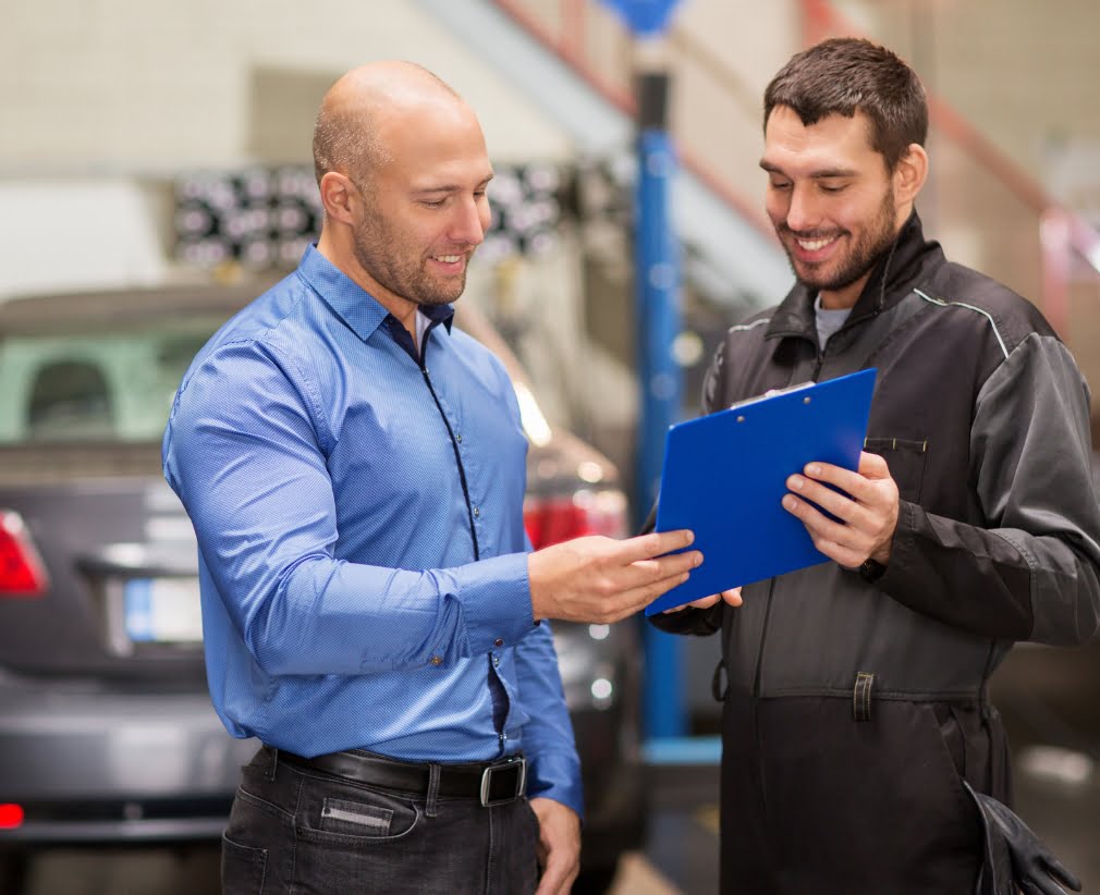 Two men stand in a garage. One, wearing a blue shirt, is pointing at a clipboard held by the other, who is wearing a black work uniform. Both are smiling, suggesting a positive interaction.