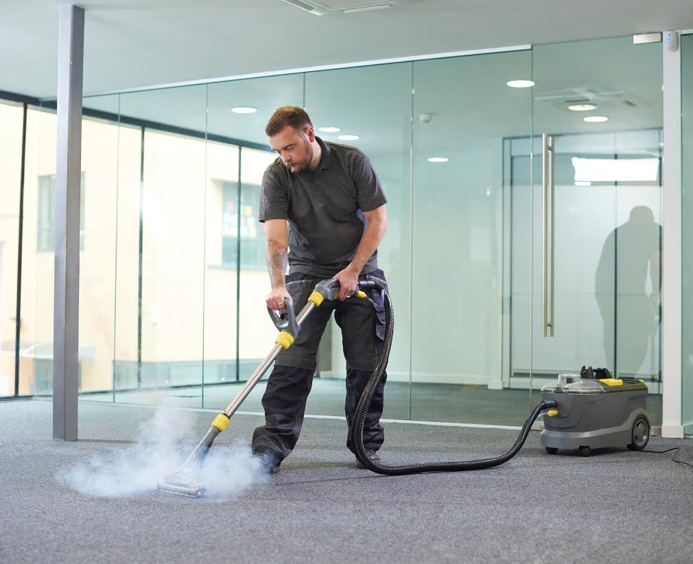 A man is using a steam cleaner to clean the carpet in an office space with glass walls.