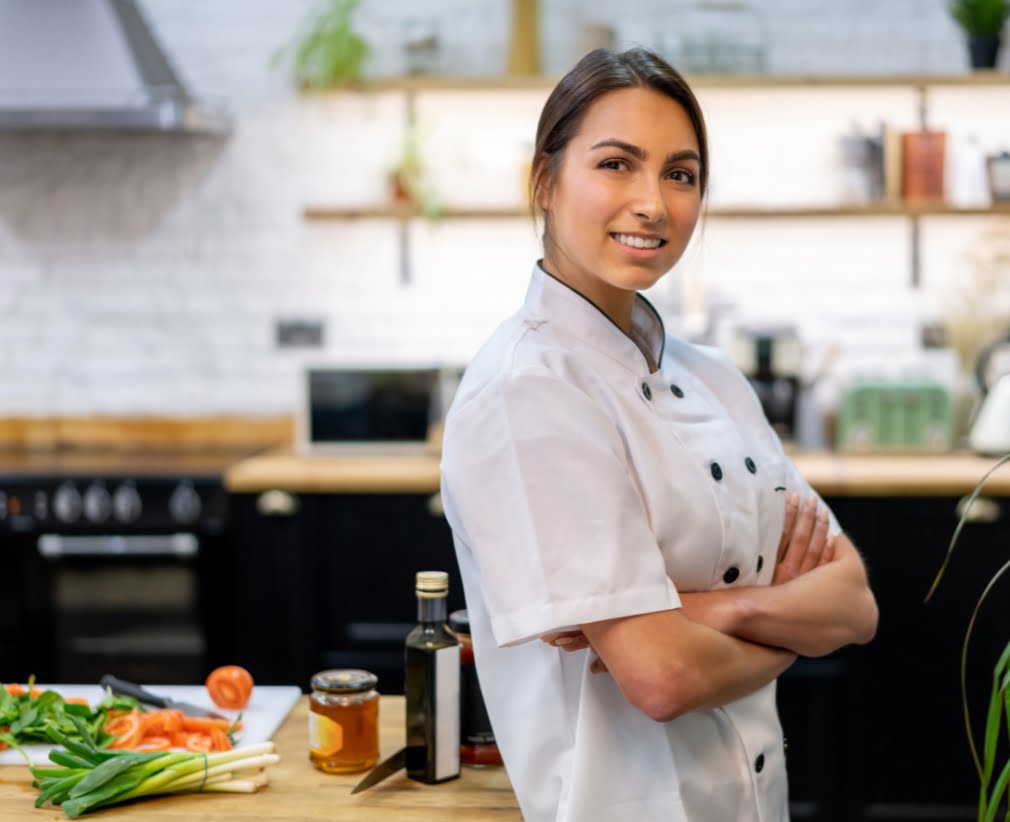 A person in a white chef's coat stands with arms crossed in a kitchen with various ingredients on the counter.