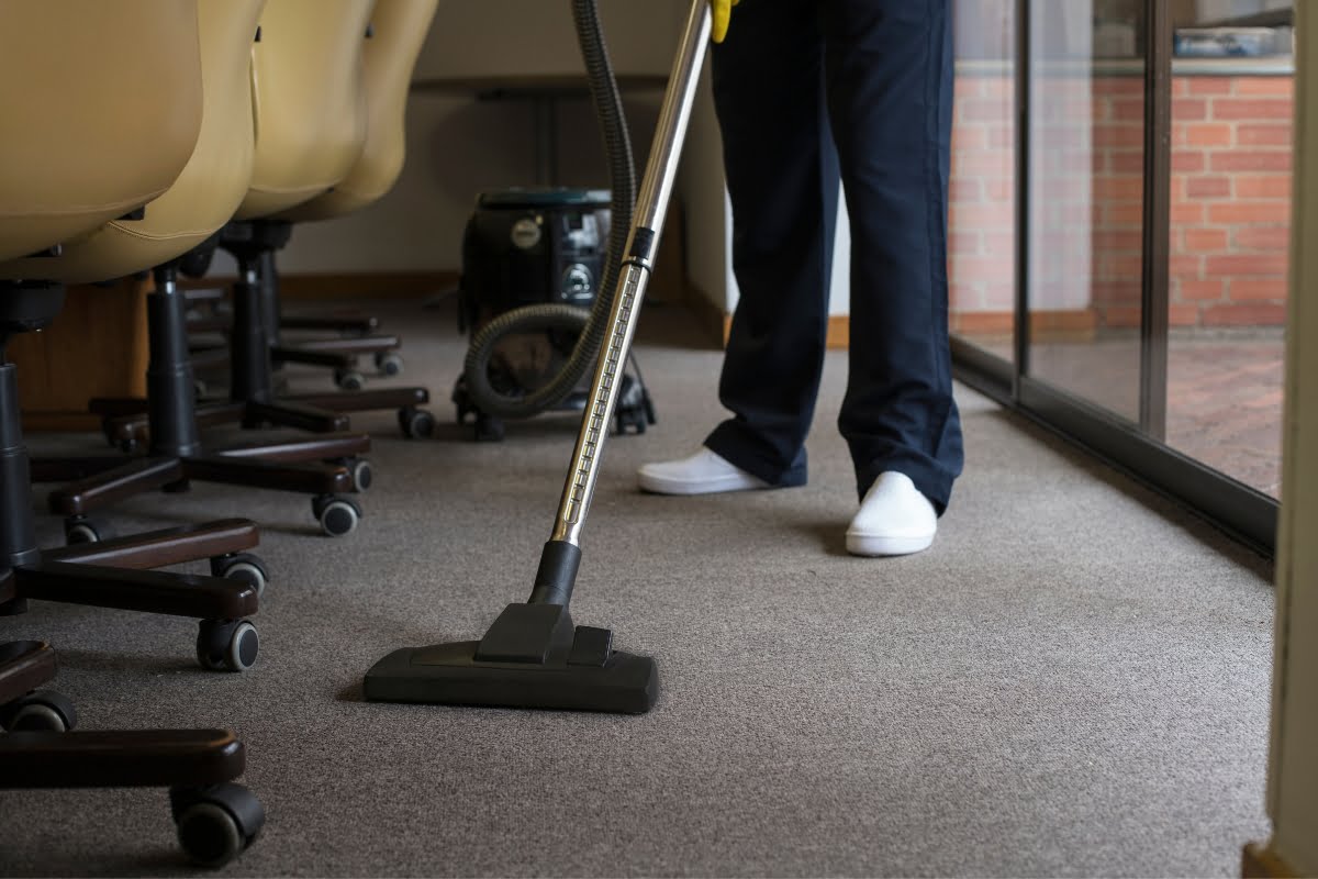 A person in dark pants and white shoes is vacuuming a carpeted floor in an office, with beige office chairs and a glass wall visible in the background.