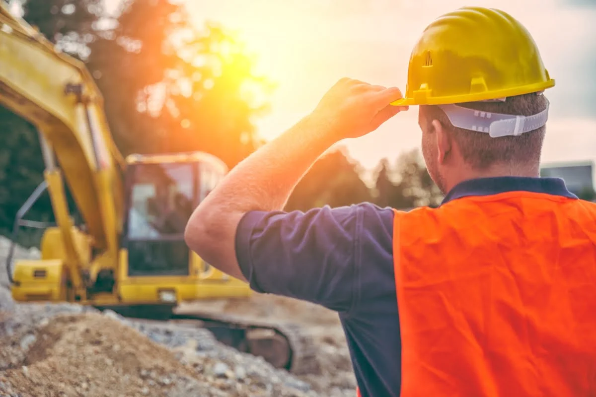 A construction worker wearing a yellow hard hat and orange vest observes an excavator at a construction site during sunset.