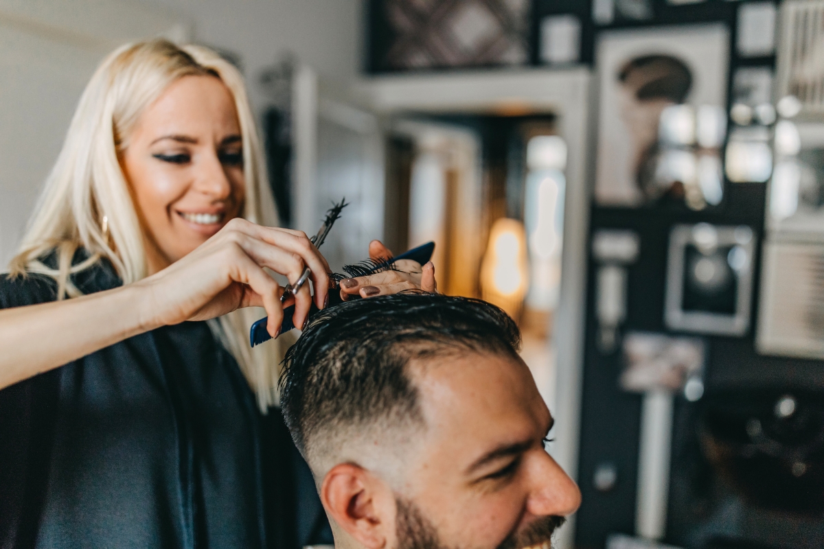 A woman with long blonde hair cuts a man's hair in a modern, well-lit salon. The man sits facing away from the camera, smiling.