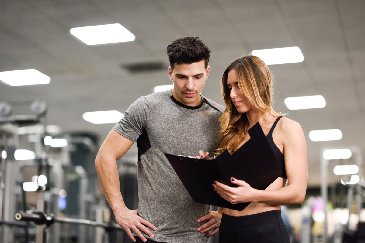 A man and personal trainer in athletic wear stand in a gym thanks to our crm for personal trainers. The woman holds a clipboard and explains something to the man. Gym equipment is visible in the background.