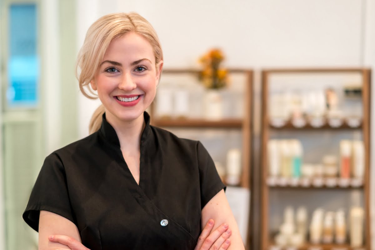 A smiling person using spa management software with blonde hair is standing with arms crossed, wearing a black shirt. Shelves with bottles and products are visible in the background.