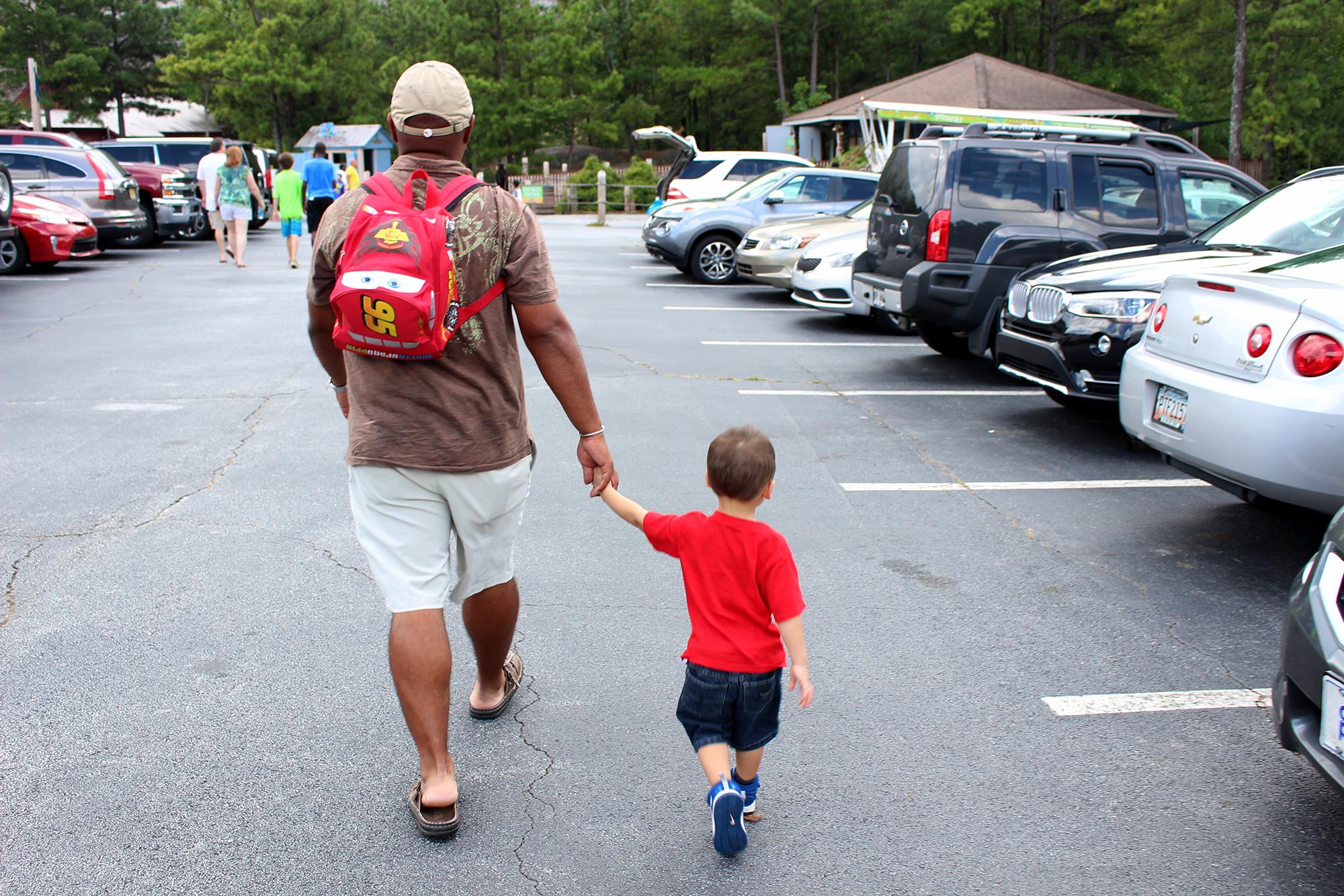 An adult and a child holding hands and walking in a parking lot with cars and trees in the background. The adult, perhaps on their way to visit theme parks in Atlanta, is wearing a hat and a red backpack, while the child sports a red shirt and denim shorts.