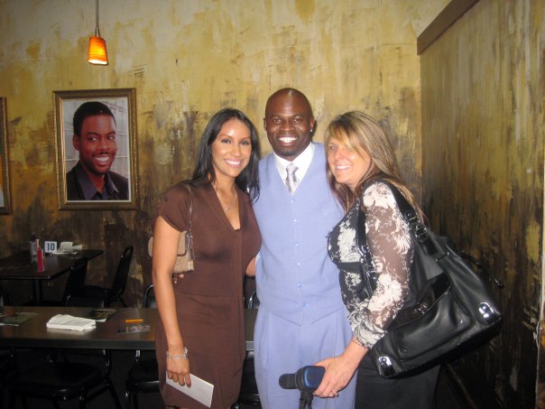 Three people posing together in a restaurant, with a framed picture of another person on the wall in the background, as if they've just left one of the nearby comedy clubs.