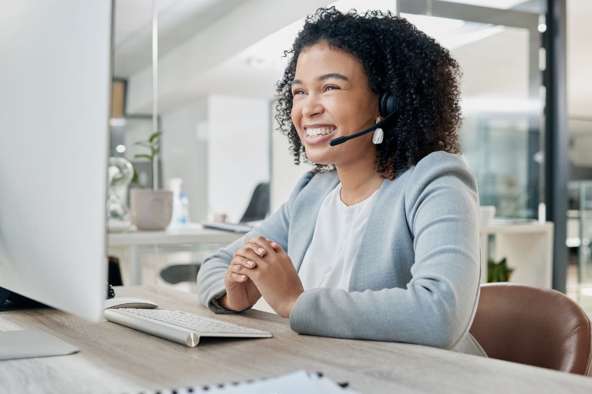 A smiling woman with curly hair wearing a headset is sitting at a desk in front of a computer monitor in a modern office, efficiently managing lead nurturing automation.