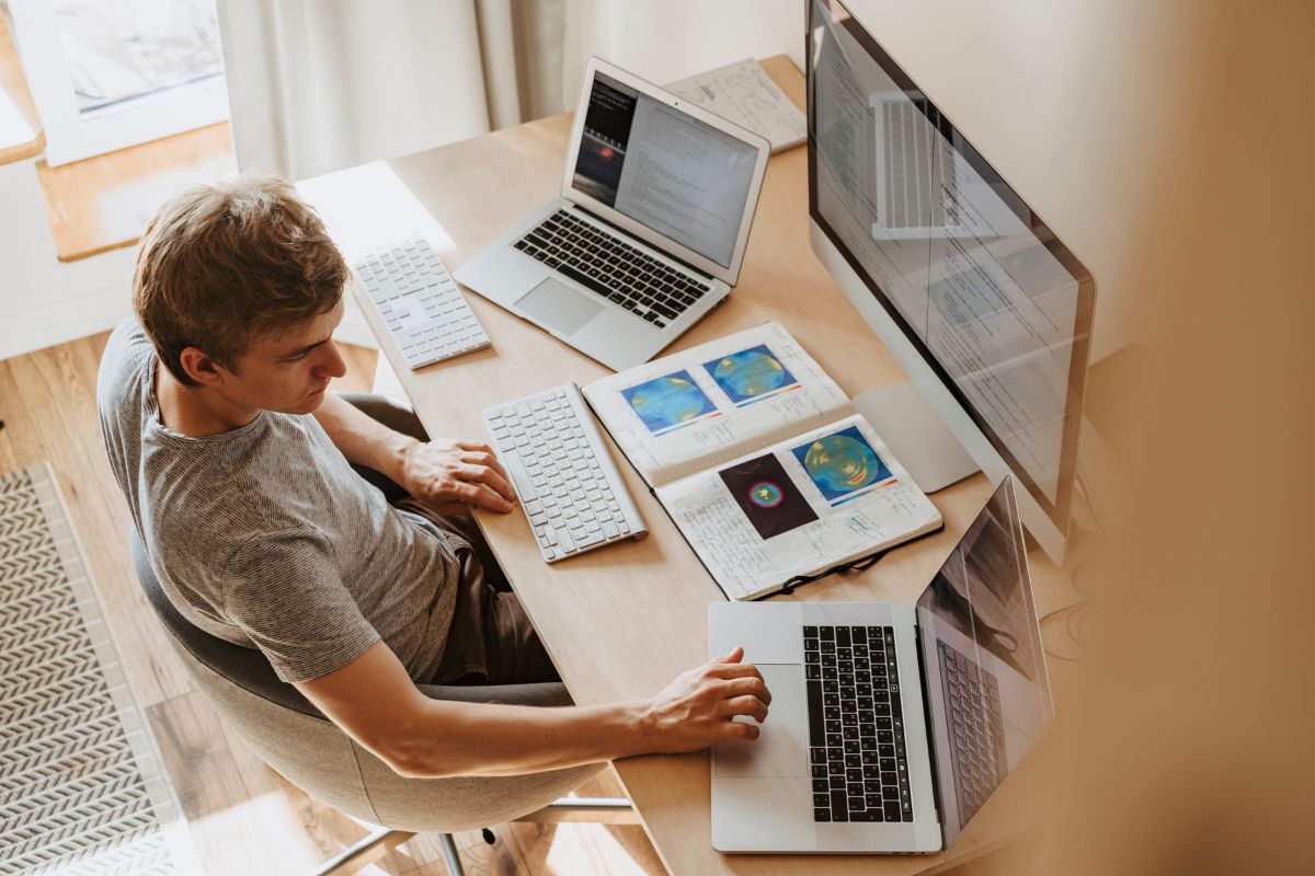 A person sits at a desk using multiple laptops and a large monitor, seamlessly managing tasks with crm integration tools, with open books and a notebook in front of them.