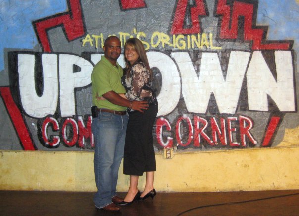 Sam and Lori Newman standing close together, smiling at the camera in front of a colorful wall that reads "UPTOWN COMEDY CORNER," one of Atlanta's premier comedy clubs.