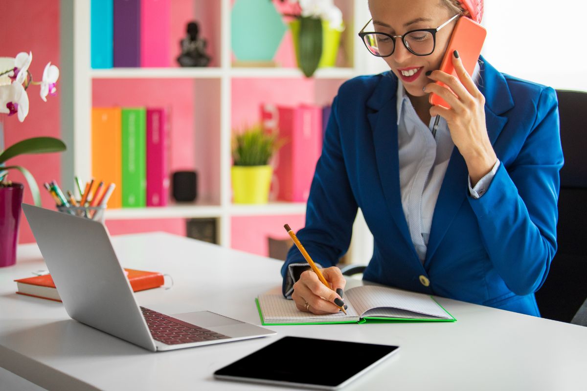 A woman in a blue blazer talks on the phone while writing in a notebook at her desk, equipped with a laptop and tablet—perhaps she's discussing ways to automate data collection.