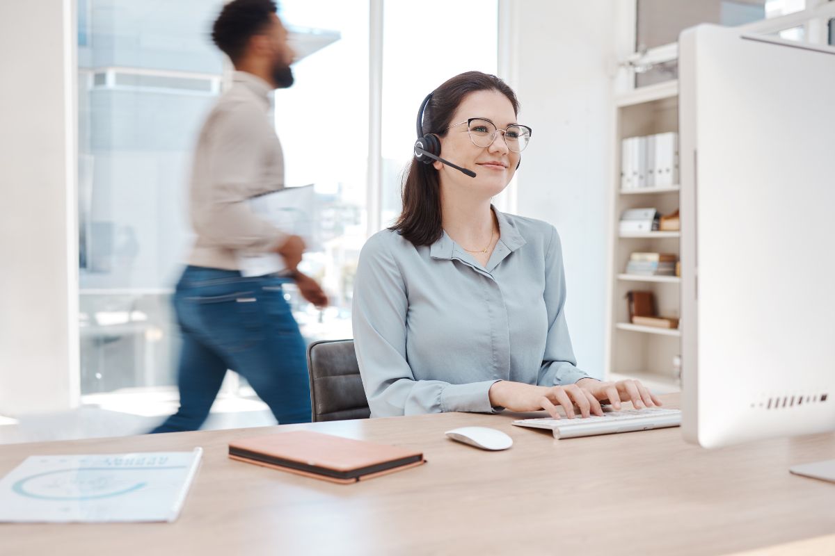 A woman with glasses wearing a headset sits at a desk typing on a keyboard, seamlessly managing CRM tasks, while a man holding a notebook walks by in the background.