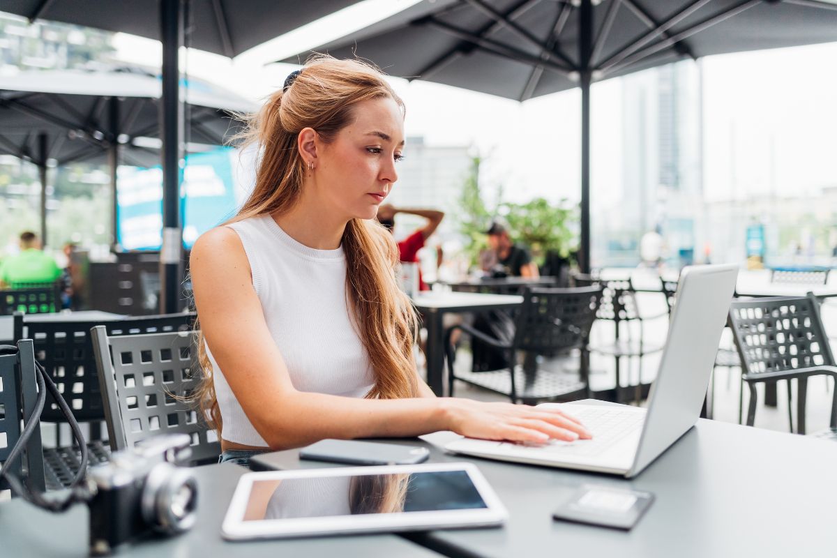 A woman with long hair sits at an outdoor café table, focused on her laptop. A camera, tablet, and phone are also on the table. Large umbrellas and city buildings provide a sophisticated backdrop as she works, perhaps researching duplicate content in SEO.