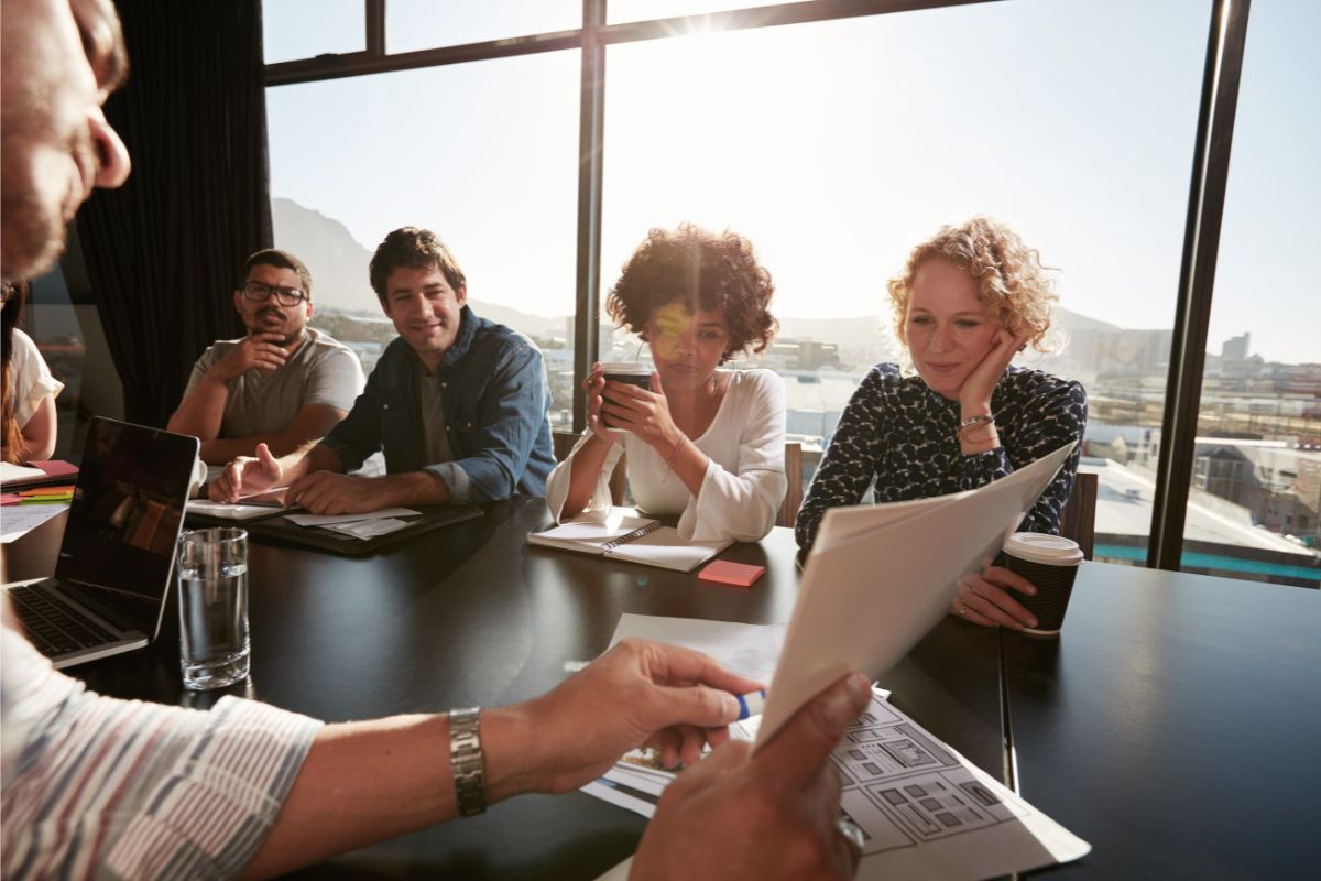 A group of people sits around a conference table, listening attentively to one individual who is holding and showing documents on lead generation. The background shows a window with a city view.