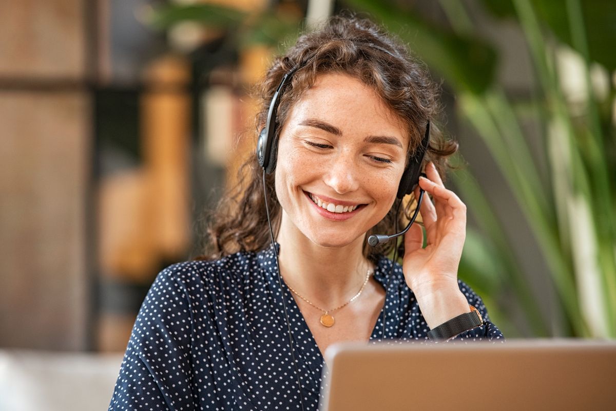 A woman wearing a headset smiles while looking at a laptop screen, focusing on lead generation marketing, with plants in the background.