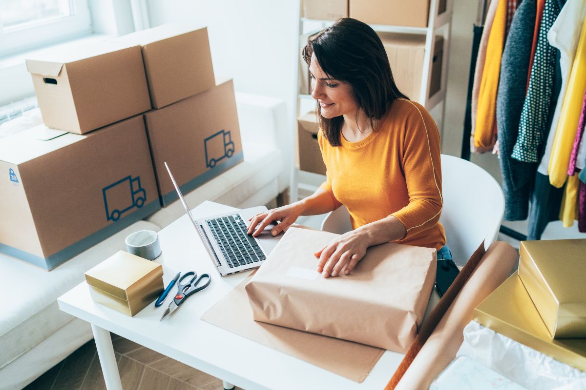 A woman in a yellow sweater sits at a desk using a laptop while packaging items, likely managing seo for multiple locations. Scissors, tape, and boxes are on the table, with more boxes stacked around her.