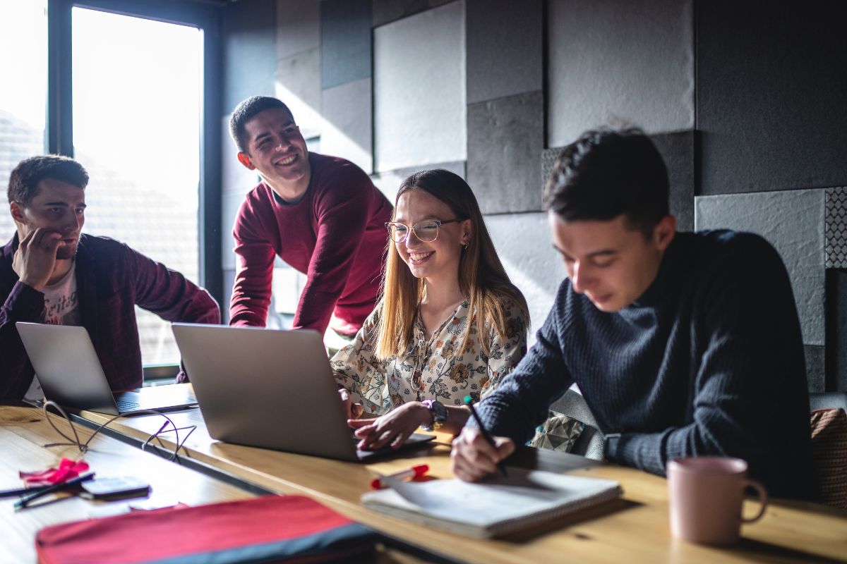 Four people are collaborating in a well-lit room, using laptops and notebooks while seated around a wooden table, discussing the implementation of CRM for business automation.