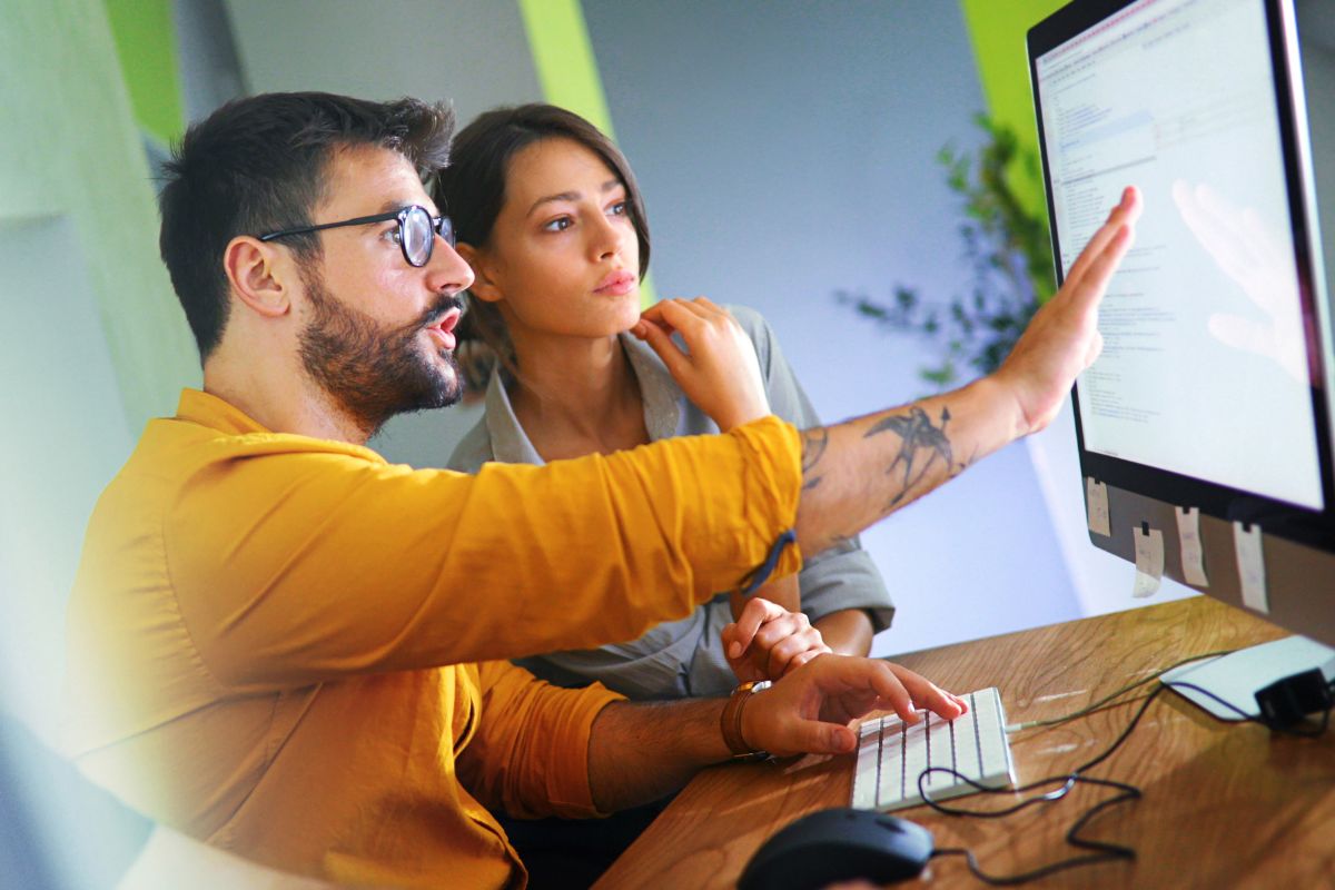 A man and woman sit at a desk looking at a computer monitor. The man points at the screen displaying CRM for business automation software, while the woman looks on attentively.