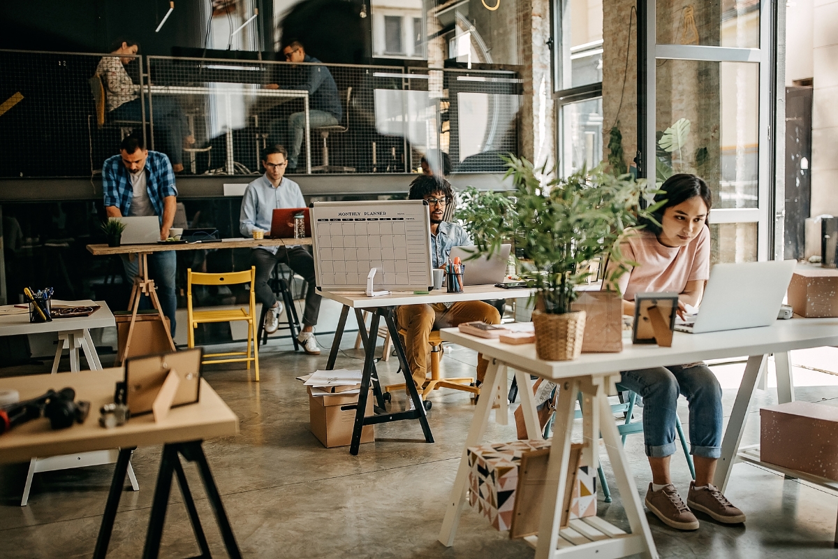 A modern office space with several people working at desks on laptops, surrounded by plants and industrial decor. Some are standing, while others are seated—an ideal setting for coworking spaces in Marietta.