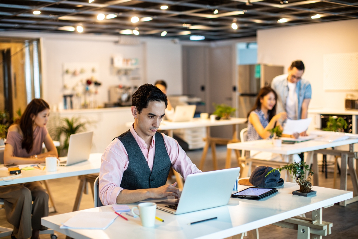 A man works on a laptop at a desk in a modern office. Other employees are working at desks in the background, reminiscent of coworking spaces in Marietta. There are plants and office supplies around the room.