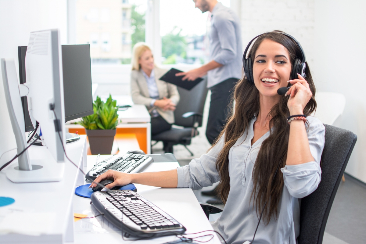 A woman wearing a headset works at a computer in an office, diligently focusing on customer data management. Two colleagues, likely discussing CRM best practices, converse in the background.