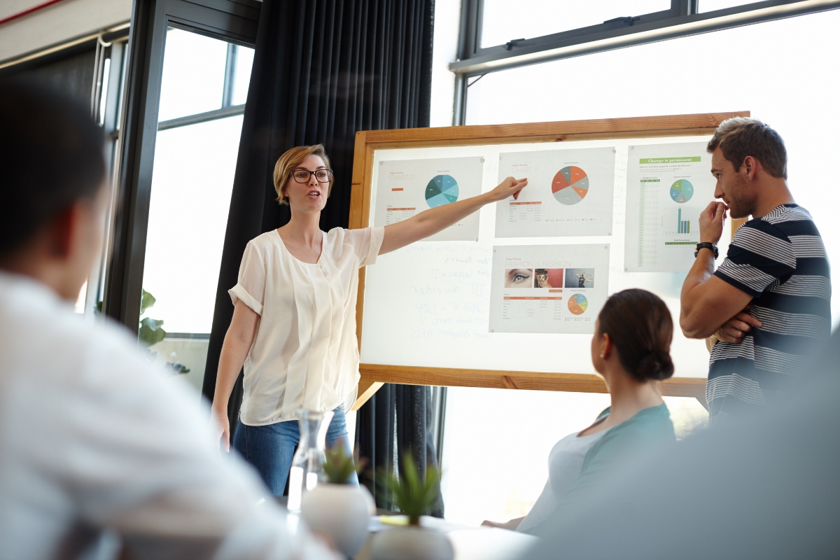 A woman in a white blouse presents charts and graphs on a whiteboard to three seated colleagues in a modern office environment, discussing CRM best practices.