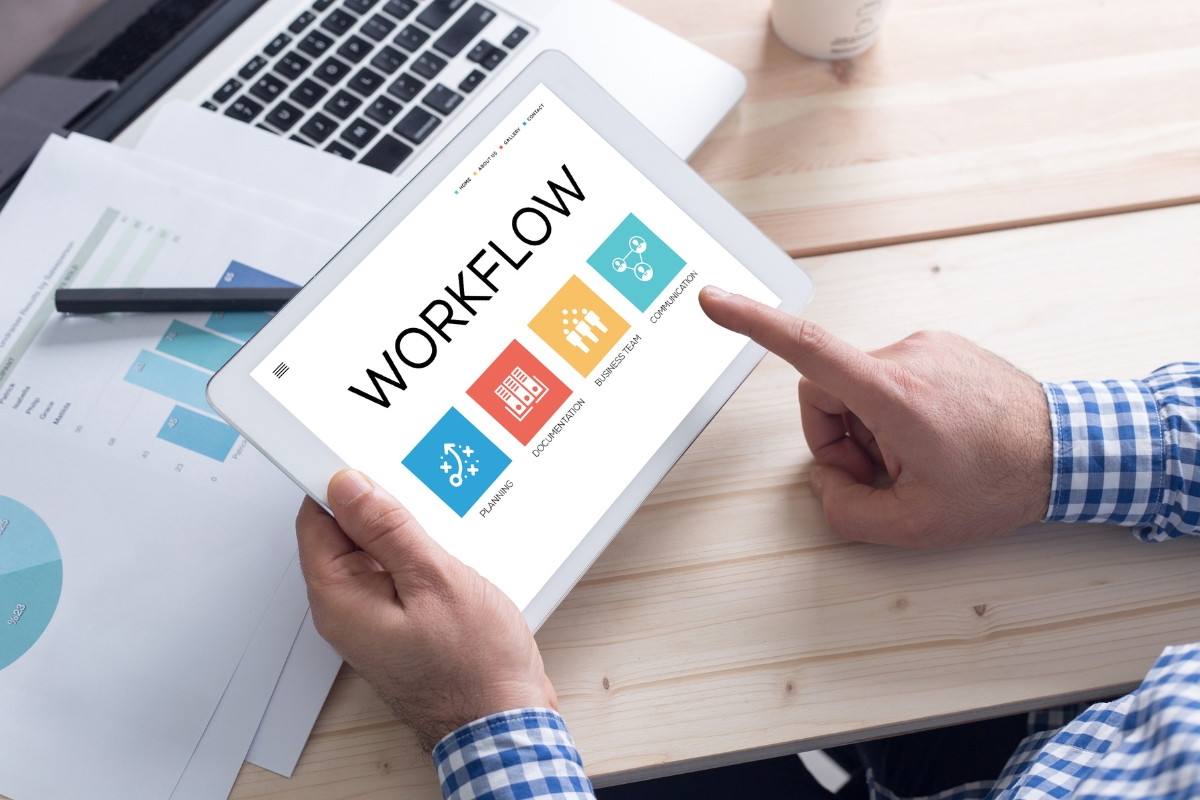 A person holding a tablet displaying a workflow screen with icons for planning, development, optimization, and communication. Beside the graphs and papers on the wooden desk, elements of customer data management are seamlessly integrated following CRM best practices.