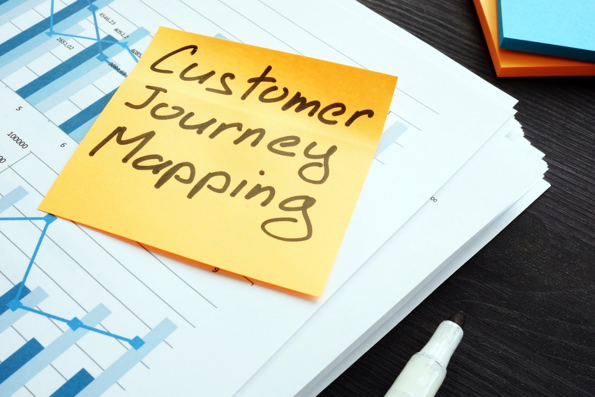 A sticky note with "Customer Journey Mapping" written on it rests atop a stack of charts and graphs, accompanied by a white pen.