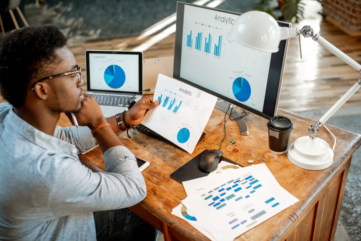 A person working at a desk with a computer, laptop, and tablet displaying analytical graphs and charts related to customer journey mapping. A coffee cup and documents with colorful charts are also on the desk.