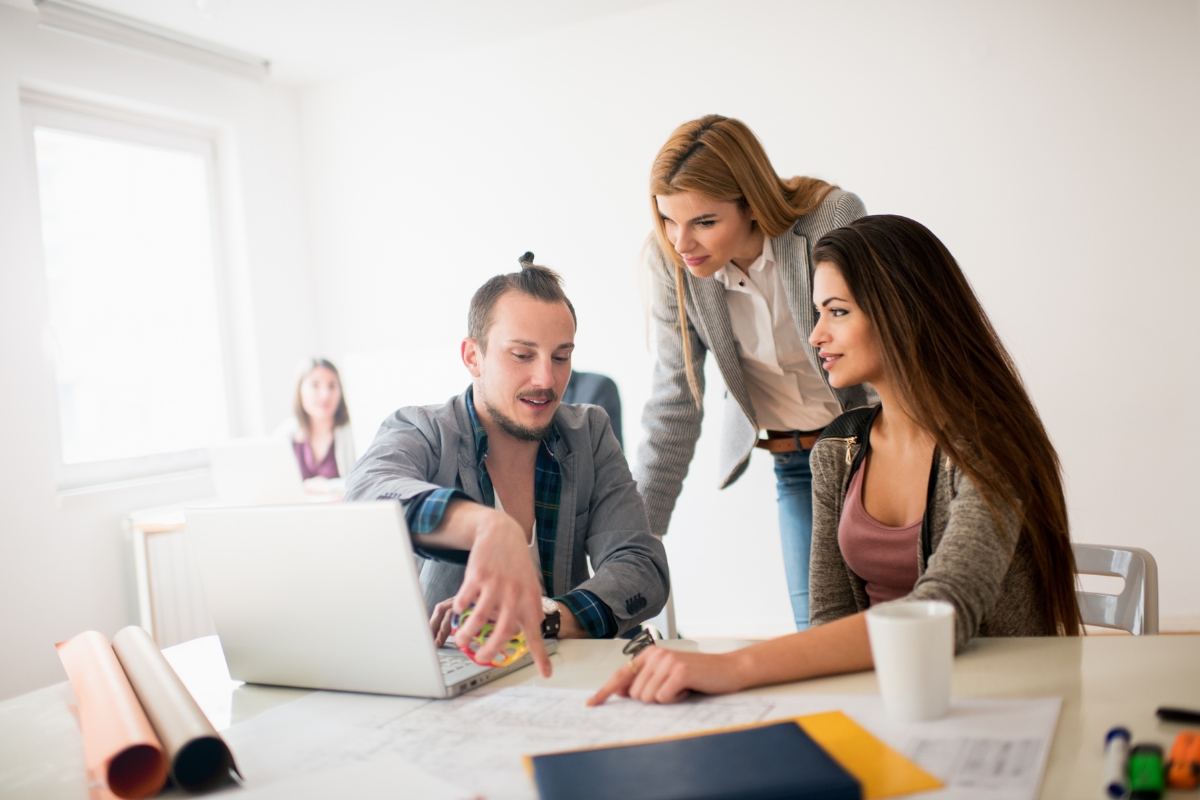 Three people are collaborating at a desk with a laptop and documents. One person points at something on the paper while another leans in to look, clearly engaging in customer journey mapping. A fourth person sits in the background, observing their progress.