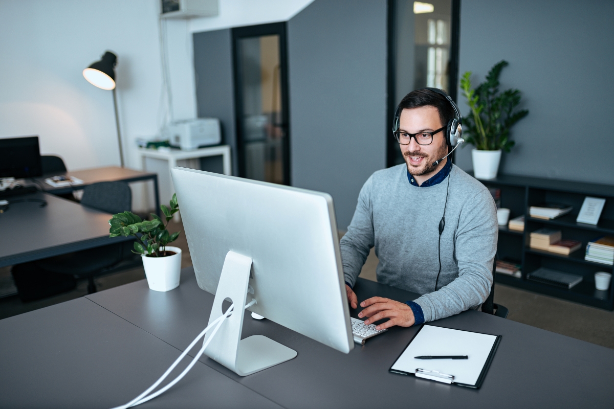 Man wearing a headset and glasses, typing on a computer at a modern office desk with a clipboard and plants nearby, perhaps deep into customer journey mapping.