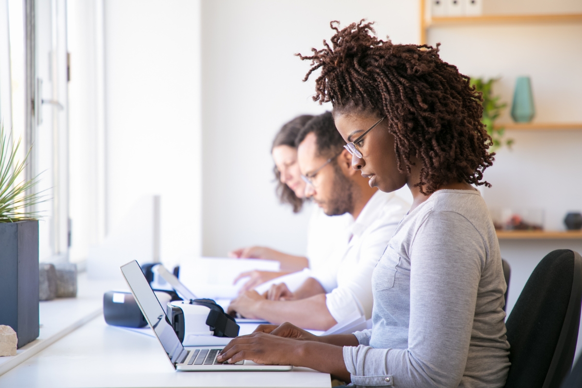 Three people are working at desks in an office, each focused on their own laptops and papers, diligently engaged in customer journey mapping. A large window provides natural light.