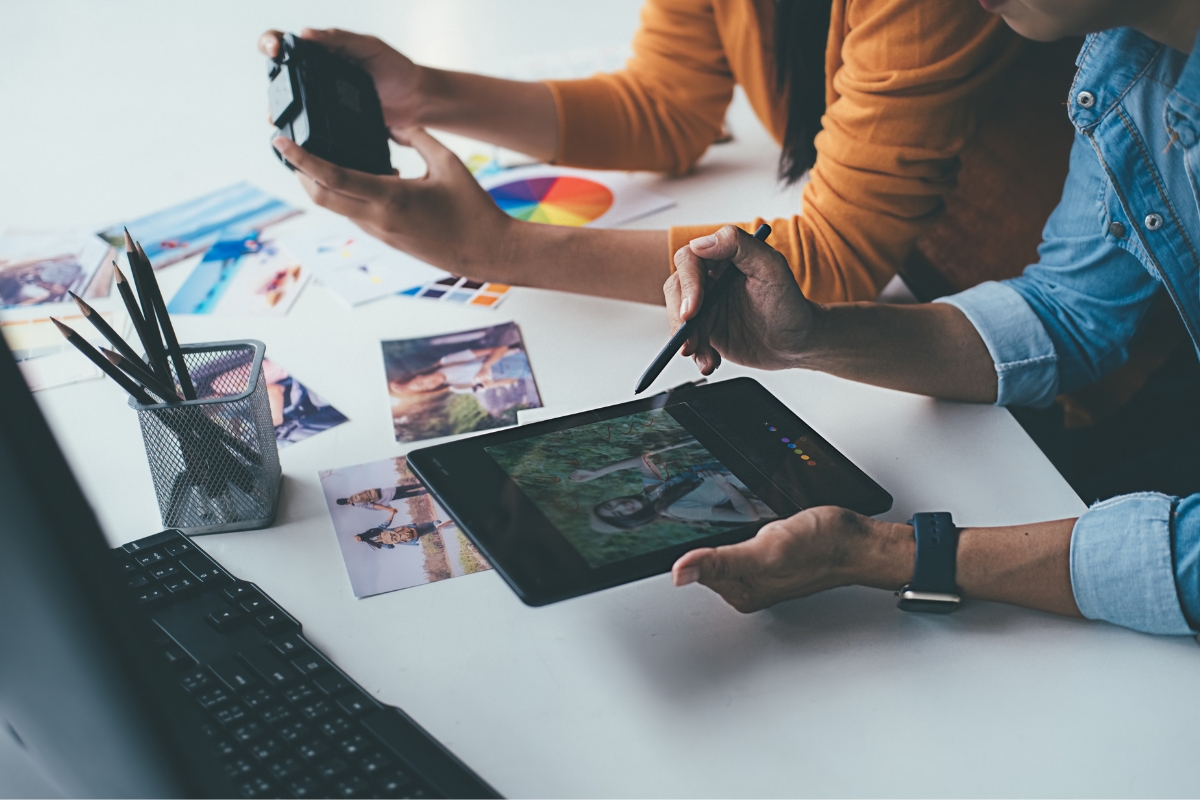 Two people collaborating on photo edits using a tablet and camera, with a pen holder and printed photos on the desk in a brightly lit room, look like they're crafting the perfect email signature marketing visuals.