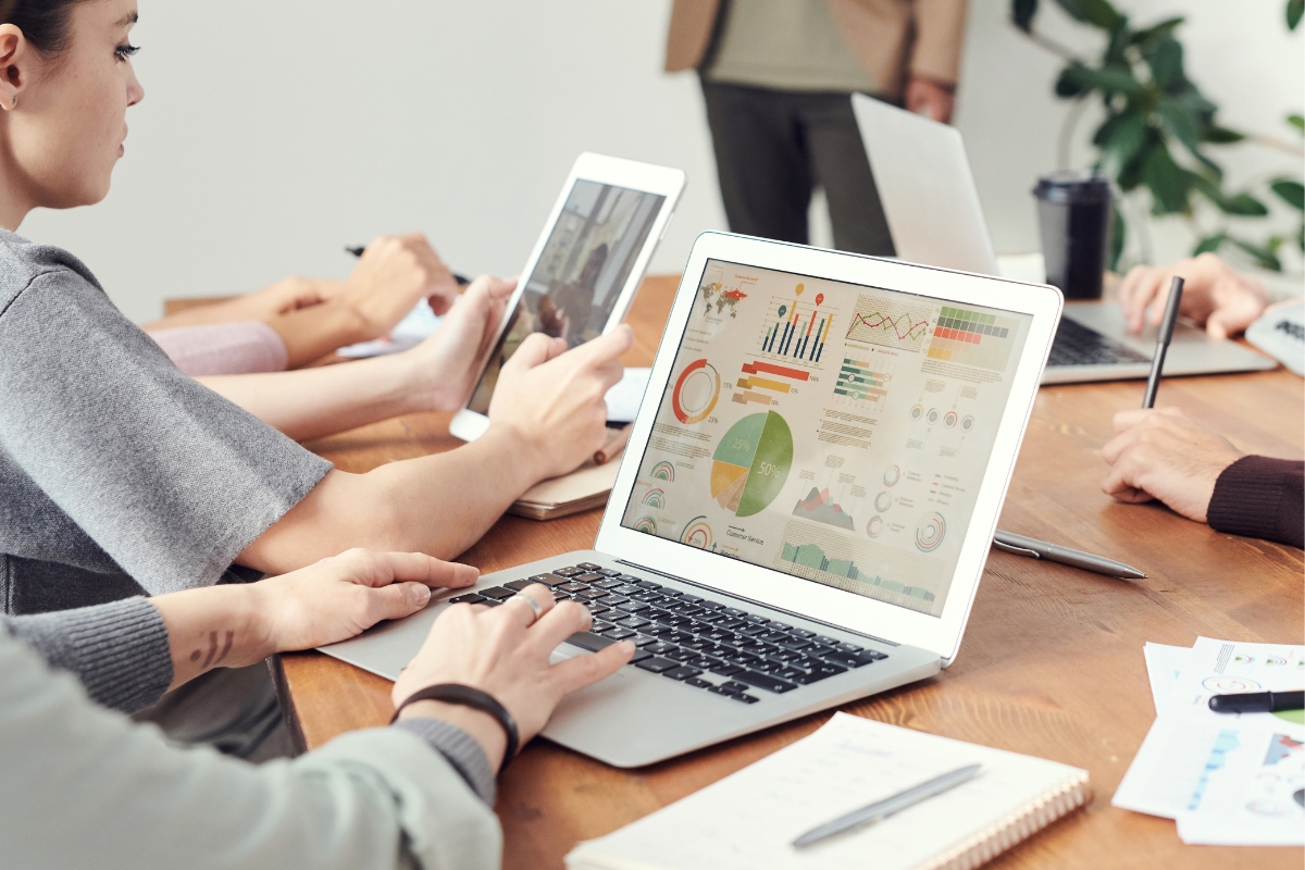 A group of individuals working at a table with laptops and tablets, focused on data and charts displayed on one of the laptop screens. Note pads and pens are also visible on the table, likely strategizing their next email signature marketing campaign.