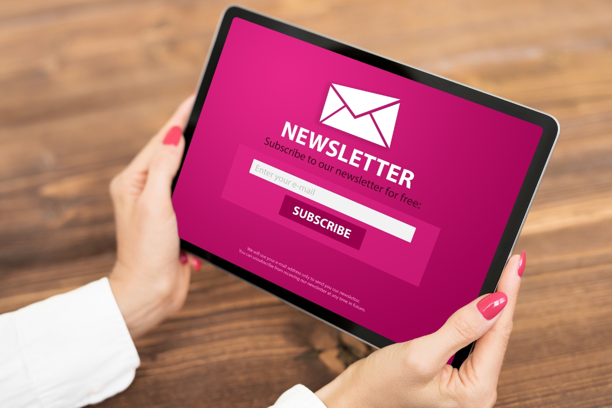 Hands holding a tablet displaying a newsletter subscription form with an email input field and a "Subscribe" button on a pink background, seamlessly blending into your email signature marketing strategy.