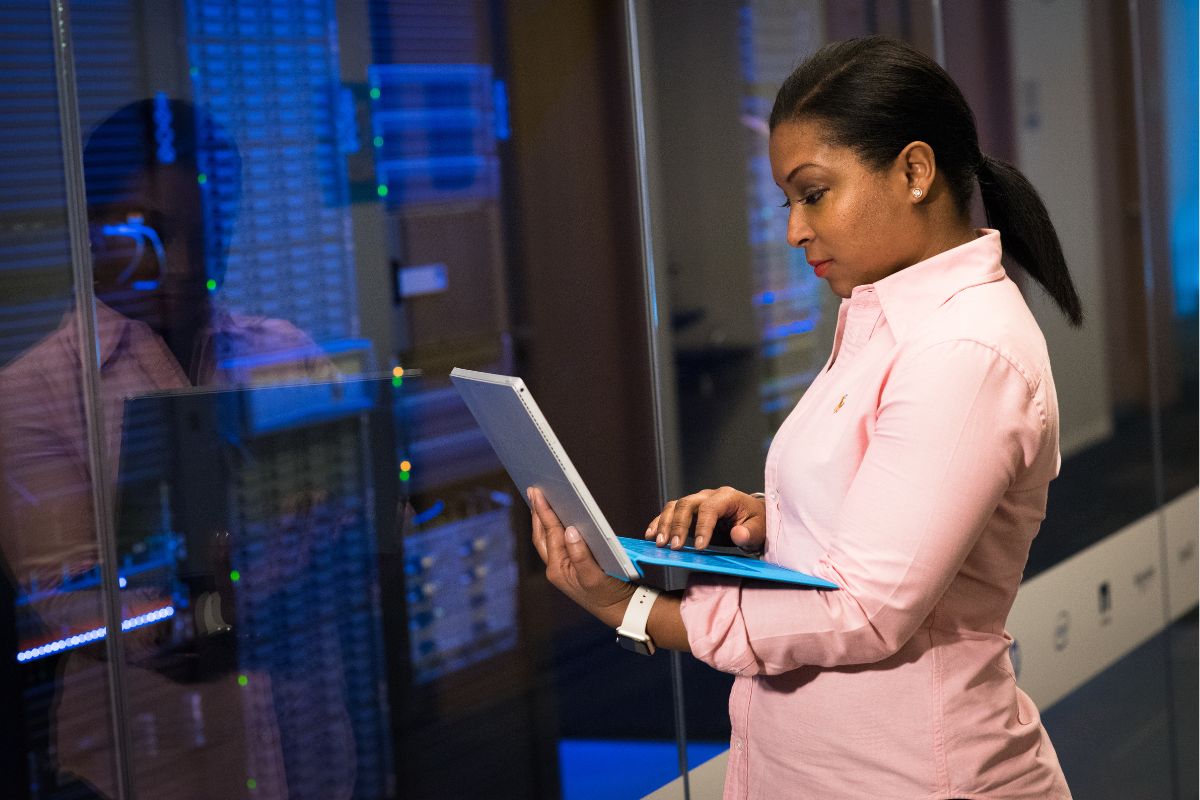 A person in a pink shirt stands in a server room holding a laptop, overseeing the implementation of affordable automation.