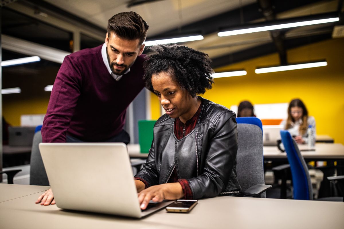 Two colleagues are collaborating at a desk on a laptop in a modern office environment. The man is standing and pointing at the screen, while the woman is sitting and typing, as they discuss workflow automation strategies.