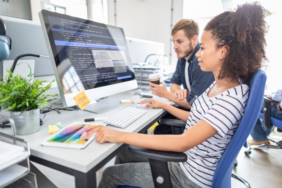 Two people working at a computer desk, one typing code on a large monitor and the other holding a coffee cup. A color palette and sticky notes are on the desk next to a small plant, while they discuss strategies for integrating CRM for business automation.