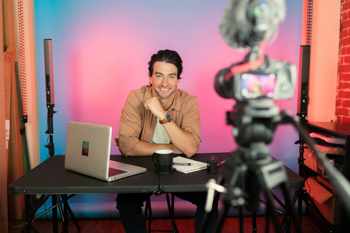 A person sits at a desk with a laptop, mug, and camera equipment, smiling at a camera set up in front of them, seamlessly blending personalized content into their marketing automation strategy.