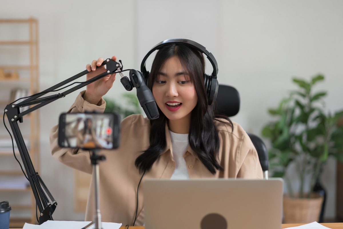 A woman wearing headphones speaks into a microphone while looking at a smartphone and laptop in a home office setting, seamlessly integrating personalized content into her marketing automation tasks.