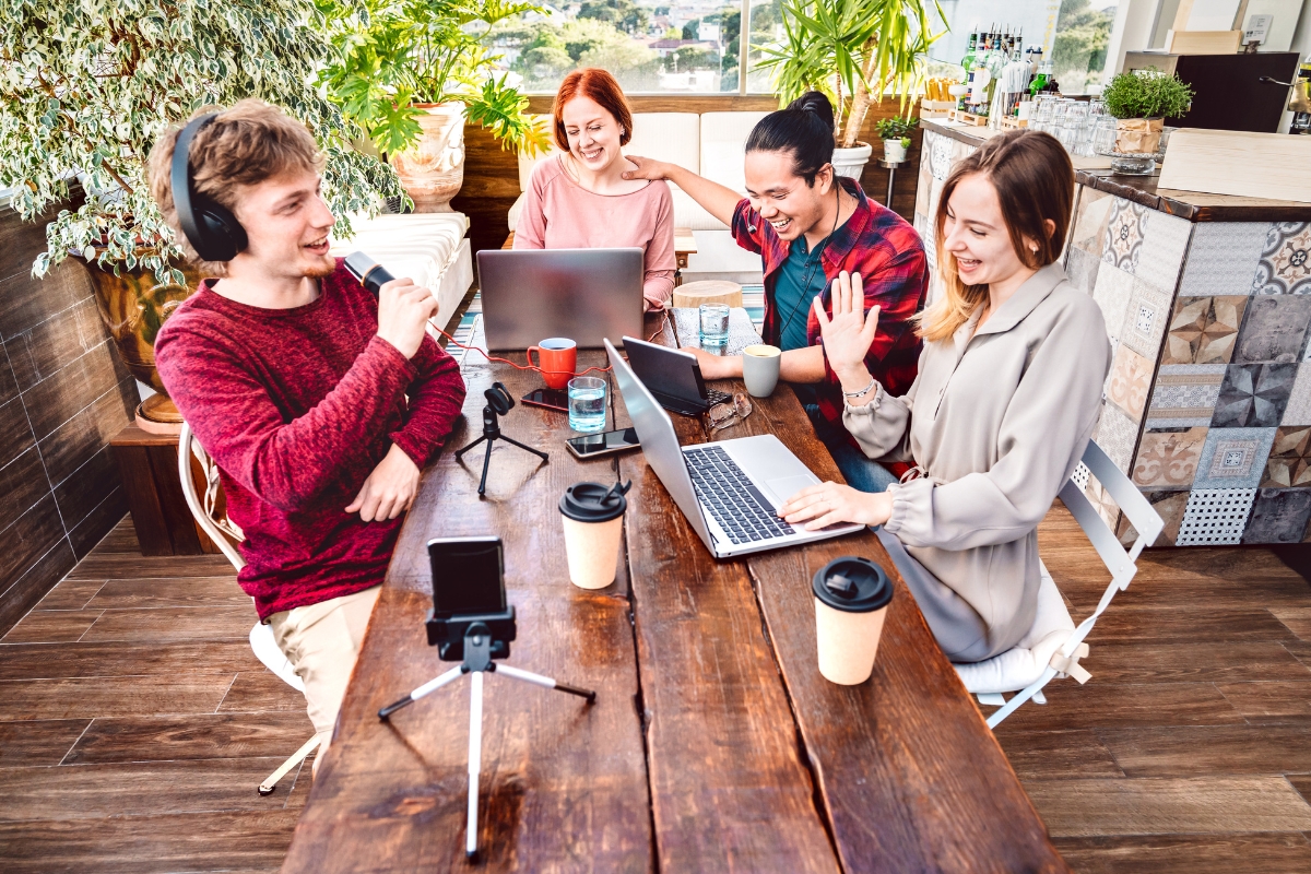 Four people sitting at a table with laptops and coffee, collaborating in a bright, casual indoor space with plants and large windows. One person, engaged in personalized content marketing automation, is wearing headphones and speaking into a microphone.