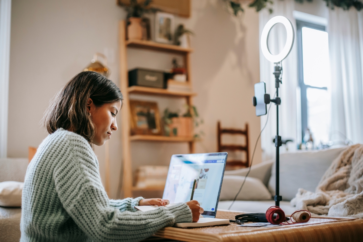 In a cozy room, a person sits at a table, writing in a notebook with a laptop in front and a ring light with phone mounted beside them, diligently working on personalized content marketing.