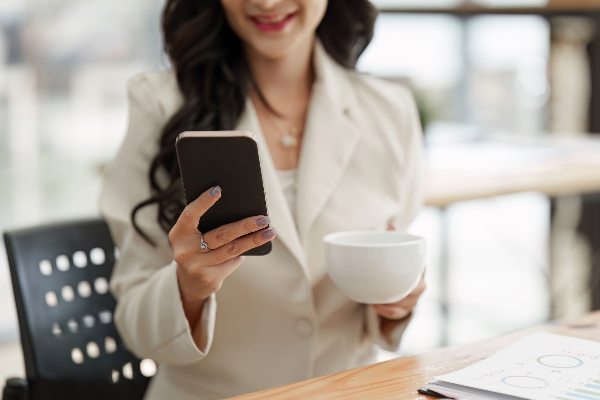 A woman in a white blazer is sitting and holding a smartphone in one hand and a coffee cup in the other. She is smiling, clearly satisfied with her sales pipeline management, as some documents lie on the table in front of her.